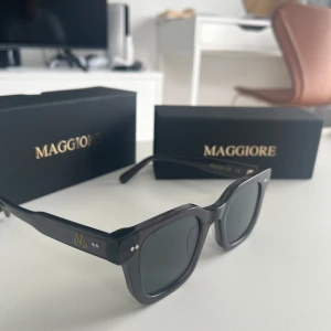 Gråa solglasögon från Maggiore - Stilrena gråa solglasögon från Maggiore med fyrkantig form och tjock båge. Snygga detaljer i guld på skalmarna och mörka glas. Perfekta för dig som vill ha en modern och clean look. Kommer med originalförpackning.