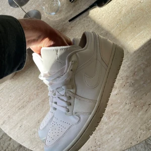 Nike Air Jordan 1 Low Triple White - Snygga Nike Air Jordan 1 Low i helvitt skinn med klassisk siluett och perforerad tå. Skorna har vit sula, snörning och ikoniska detaljer som Swoosh-loggan och Air Jordan Wings på hälen. Perfekta för dig som gillar clean och stilren streetwear.