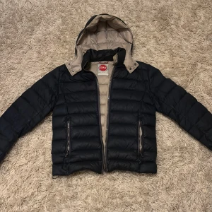 Mörkblå dunjacka från Colmar - Snygg mörkblå dunjacka från Colmar med beige detaljer och avtagbar huva. Jackan har två dragkedjeförsedda fickor och quiltad design som ger en modern look. Perfekt för kalla dagar och enkel att matcha med olika outfits. Skriv om frågor! Tar gärna emot prisförslag!