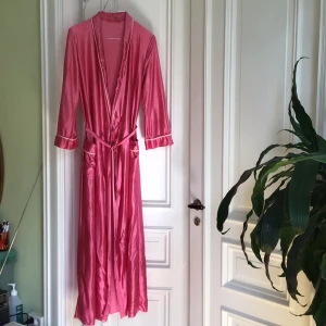 Lång rosa sidenklänning med bälte - Säljer en tunn lång morgonrock i skimrande rosa siden känsla. Den  har långa ärmar och fina ljusa kantdetaljer vid krage och ärmslut. Perfekt för dig på resor som tar lite plats i resväskan .  Mjuk material och en lyxig känsla.