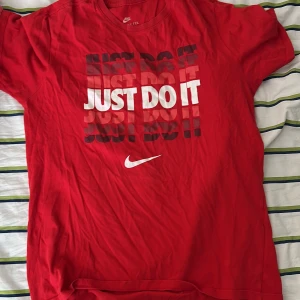 Röd Nike Just Do It t-shirt. Storlek M vuxna.  - Röd t-shirt från Nike med det klassiska 'Just Do It'-trycket i vitt och svart framtill. T-shirten har rund halsringning och korta ärmar. Tillverkad i mjuk bomull, perfekt för en sportig och avslappnad stil.