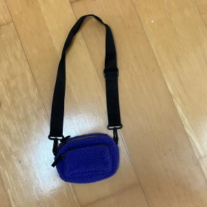 Lila fleece axelväska med dragkedja - En liten och smidig axelväska i lila fleece med svart justerbart band. Väskan har ett huvudfack och en mindre ficka framtill, båda med dragkedja. Perfekt för att bära det viktigaste och ger en cool vibe till din outfit.