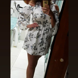 Vit blommig kortklänning med offshoulder - Supersöt vit kortklänning med blommigt mönster i blått och svart. Klänningen har offshoulder och vida trekvartsärmar, samt en smockad midja som ger en snygg siluett. Perfekt för varma dagar och har en lätt och luftig känsla.är en fast använd en gång. 