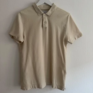 Beige pikétröja från These Glory Days - Snygg beige pikétröja från These Glory Days i mjuk bomull. Klassisk krage med två knappar framtill och korta ärmar. Perfekt för en chill och clean look. Passar dig som gillar stilrena och enkla plagg. *Liten fläck längst ned, går troligvis bort med lite kärlek*