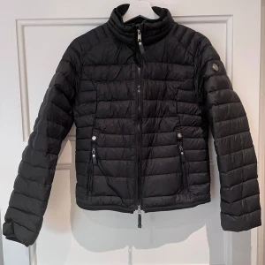 Svart pufferjacka från Cedrico - Snygg svart pufferjacka från Cedrico i storlek M. Jackan har quiltad design, hög krage och två dragkedjeförsedda fickor framtill. Stilren look med diskret logga på ärmen.