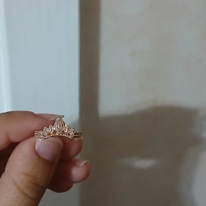 Guldig kronformad ring med stenar - Superfin ring i guldton roseguld med en kronformad design och små gnistrande stenar typ framtill. Ringen har detaljerade kanter som ger en lyxig känsla och passar perfekt för dig som gillar unika smycken med lite extra bling.