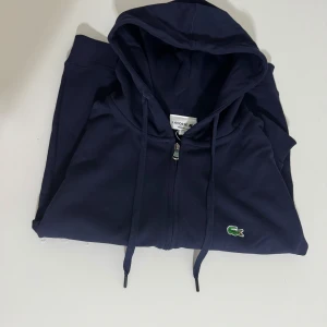 Mörkblå hoodie från Lacoste - Snygg mörkblå hoodie från Lacoste med dragkedja och klassisk krokodillogga på bröstet. Tröjan har huva med snörning, känguruficka och ribbade muddar. Perfekt för chill dagar och streetstyle. Tillverkad i mjuk bomull för skön känsla.