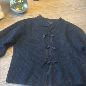 Svart stickad kofta med rosetter - Mysig svart stickad kofta med lång ärm och fyra dekorativa rosetter framtill istället för knappar. Koftan har en rak passform och är perfekt att slänga över en topp för en cool och söt look.