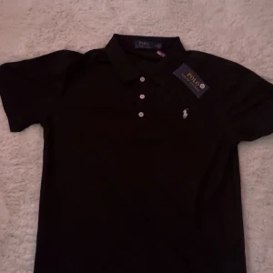 Svart Polo Ralph Lauren t-shirt M - Svart t-shirt från Polo Ralph Lauren i klassisk pikémodell med krage och tre vita knappar framtill. Ikoniska broderade loggan i vitt på bröstet. T-shirten är kortärmad och tillverkad i mjuk bomull som känns skön mot huden.