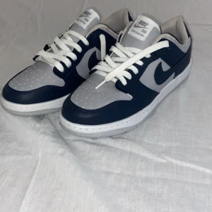 Nike SB Dunk Low marinblå/grå 44 - Nike SB Dunk Low sneakers i marinblått och grått med vit snörning och klassisk rund tå. Ovandel i skinn och syntet, perforerad tåbox och vit yttersula med grått mönster. Snygg kontrast mellan färgerna och tydlig Nike-logga på hälen.