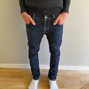 Mörkblå slim jeans från Nudie Jeans - Säljer ett par mörkblå jeans från Nudie Jeans med snygga kontrastsömmar och klassisk broderad bakficka. Modellen har normal passform och raka ben, tillverkade i slitstarkt bomullsmaterial. Perfekt för dig som gillar stilrena och tidlösa jeans.