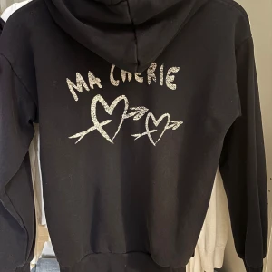 Ma Chérie Gina Tricot - Svart hoodie från Gina Tricot med texten 'Ma Chérie' och två hjärtan med pilar i vitt tryck på ryggen. Tröjan har huva, snörning och ribbade muddar. Perfekt för dig som gillar coola tryck och mjuka material.