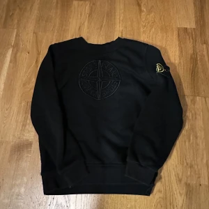 Svart Stone Island sweatshirt - Svart sweatshirt från Stone Island med stort broderat logomärke på bröstet och klassisk patch på vänster ärm. Tröjan har rund hals och långa ärmar, tillverkad i mjuk bomull som är skön att ha på sig. Perfekt för dig som gillar streetwear och vill ha något stilrent.