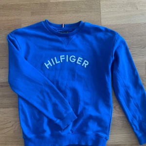 Blå Tommy Hilfiger sweatshirt - Snygg blå sweatshirt från Tommy Hilfiger med vit logga framtill. Tröjan har rund hals, ribbade muddar och klassisk passform. Perfekt för dig som gillar stilrena och sportiga plagg med ikonisk branding.