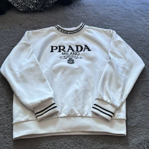 Vit sweatshirt från Prada med brodyr - Vit sweatshirt från Prada med svart broderad logga och text på bröstet. Tröjan har svarta ränder på muddar och krage samt rund halsringning med broderad Prada-text. Långärmad och i mjukt bomullsmaterial, perfekt för en clean och stilren look.