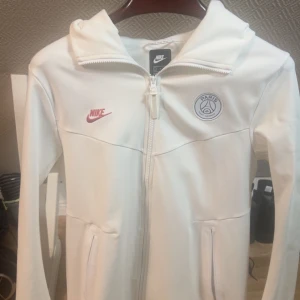 Vit Nike PSG vindjacka med huva - Snygg vit vindjacka från Nike med PSG-logga på bröstet och röd Nike-swoosh. Jackan har huva, hel dragkedja och två fickor med dragkedja. Perfekt för dig som gillar sportig stil och vill representera Paris Saint-Germain.