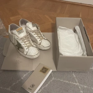 Golden Goose Superstar sneakers vit/oliv - Golden Goose Superstar sneakers i vitt skinn och textil med olivgrön stjärna på sidan. Klassisk rund tå, beige snörning och perforerade detaljer framtill. Snygg vintagekänsla och ikonisk design, perfekt för dig som gillar streetstyle. Äkthetsbevis och box medföljer. Är lite slitna i hälen på undersidan av skon.