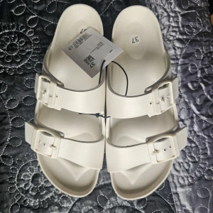 Beiga sandaler med dubbla spännen - Snygga beiga sandaler med två breda remmar och justerbara spännen. Platt sula och öppen tå gör dem perfekta för varma dagar. Tillverkade i syntetmaterial och passar dig som gillar enkel och stilren design.