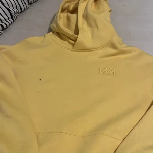 Gul hoodie från WA med broderad logga - Snygg ljusgul hoodie från WA med broderad logga på bröstet och en patch nertill. Tröjan har luva och långa ärmar. Materialet är mjuk bomull som känns skönt mot huden.