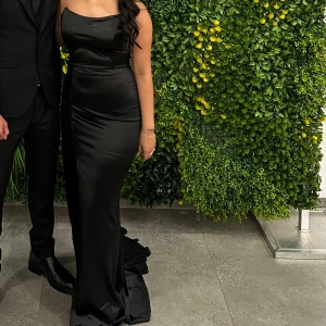 Svart långklänning med slits - Supersnygg svart långklänning i satinliknande material med figurnära passform. Klänningen är axelbandslös och har en hög slits på ena sidan och kor litet släp tyg på andra sidan. Perfekt till fest eller bröllop 