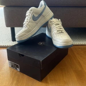 Nike Air Force 1 Summit White/Blue - Nike Air Force 1 LXX i färgen Summit White och University Blue. Skorna har en vit ovandel i skinnliknande material med ljusblå mockadetaljer på swoosh och häl, samt matchande blå yttersula. Klassisk låg sneaker med snörning och rund tå. Perfekt för dig som gillar stilrena och fräscha sneakers.