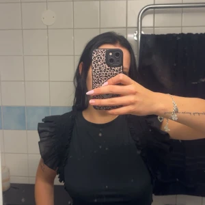 Svart blus med volangärmar från Misu - Snygg svart blus från Misu i storlek M. Toppen har coola volangärmar med rynkade detaljer som ger extra attityd. Materialet är mjukt och följsamt, perfekt för dig som gillar att sticka ut med din stil. Rund halsringning och figurnära passform.