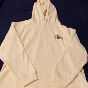 Stüssy hoodie i ljus beige, L - Säljer en clean hoodie från Stüssy i ljus beige med svart logga på bröstet. Tröjan har huva med snörning och ribbade muddar. Perfekt för chill dagar och streetwear-stil. Materialet är mjuk bomull och passformen är normal.