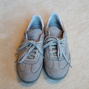 Grå adidas sneakers i mocka strl 40 - Snygga grå adidas sneakers i mocka med klassisk låg siluett och rund tå. Skorna har snörning, diskret adidas-logo på plösen och sidan samt en mörkgrå sula. Perfekta för dig som gillar stilrena och cleana sneakers.