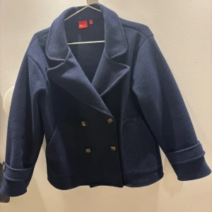 Mörkblå kort kappa från RYVLS - Snygg, marinblå, kort, kappa från RYVLS med dubbelknäppning . Kappa har två framfickor, dekorativa knappar och är tillverkad i ett mjukt ullmaterial. Storlek 158/164, passar mig med storlek xs/s.