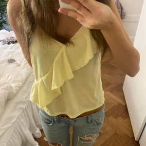 Gul linnetopp med volang - Söt ljusgul linnetopp med volangdetalj framtill och v-ringad hals. Tunn och luftig, perfekt för varma dagar. Ärmlös modell med smala axelband och avslappnad passform. Superfin till jeans eller kjol för en fräsch sommarlook! 💛💛
