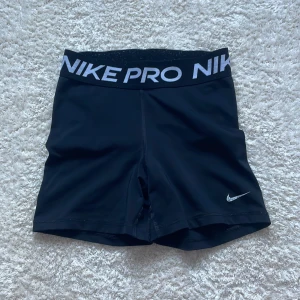 Nike Pro svarta träningstights-shorts - Svarta Nike Pro shorts i tight modell med bred resår i midjan där det står NIKE PRO i vitt. Sitter skönt och följsamt, perfekta för träning eller sport. Materialet är stretchigt och andas, med klassisk Nike Swoosh-logga på benet. Ett jättelitet hål i grenen, inget som man ser när man har på sig dem och hålet är inte rakt igenom bara i ”ytterlagret”💞