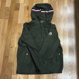Grön moncler windbreaker  - Snygg mörkgrön vindjacka från Moncler med huva och dragkedja. Jackan har Moncler-logga på bröstet och coola ränder i rött, vitt och blått på huvan. Tillverkad i polyester och perfekt för dig som gillar stilren och sportig look. Size 2 helt ny nästan använd 3 gånger