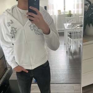 Vit hoodie med strasstenar och BLING-tryck - Säljer en vit hoodie med dragkedja och huva. På bröstet finns ett motiv av en ängel med pilbåge i strasstenar samt texten 'BLING' i samma stil. Hoodien har fickor framtill och är i mjukt material.