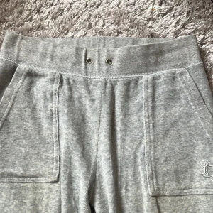 Grå juicy couture mjukisbyxor med fickor - Säljer ett par grå juicy couture mjukisbyxor med bred resår i midjan och stora fickor både fram och bak. Byxorna har en avslappnad passform och är perfekta för chill dagar. Liten broderad detalj på ena fickan framtill. (Saknar bandet i midjan)