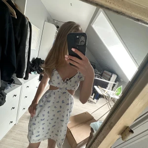 Vit blommig klänning - Söt vit klänning med blått blommönster, cut-out i midjan och rosett framtill. Klänningen har korta ärmar och är i en lätt, somrig modell. Pris kan diskuteras💞