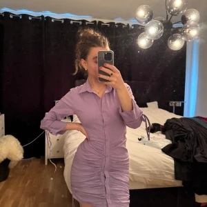Ljuslila skjortklänning från Fashion Nova - Säljer en ljuslila skjortklänning från Fashion Nova med knappar framtill och snörningsdetaljer vid ärmslut och sidor. Klänningen har krage och är figurnära med rynkade partier längs sidorna. Perfekt för dig som gillar trendiga och feminina plagg.