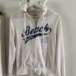 Vit hoodie med blå text från H&M - Vit hoodie med dragkedja från H&M, storlek 36/38. Tröjan har blå text med 'Beach Surf Co.' framtill, huva med dragsko och fickor. Perfekt för en avslappnad stil. Använd lite fläckig annars bra strl S