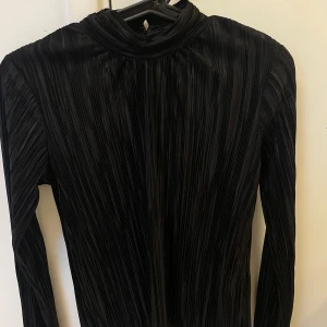 Svart plisserad långärmad blus från i Gina Tricot - Säljer en svart plisserad blus från Gina Tricot med hög hals och långa ärmar. Blusen har en elegant struktur och stängs med knapp i nacken. Perfekt för dig som gillar stilrena och trendiga plagg.