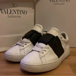 Valentino Garavani sneakers med svart detalj - Säljer ett par vita sneakers från Valentino Garavani med en bred svart läderdetalj över mitten. Klassisk rund tå och platt sula. Skorna har en stilren och modern design, perfekta för dig som gillar exklusiva märken. Dom är tyvärr lite slitna men inget man märker när man tittar på dom