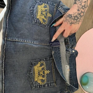 Mörkblå jeanskjol med guldbroderade kronor - Säljer en mörkblå jeanskjol från Victoria Beckham med två bakfickor dekorerade med stora guldbroderade kronor. Kjolen har klassisk jeanslook och knappstängning framtill. Perfekt för dig som vill sticka ut med detaljer! Säljes för 600 kr