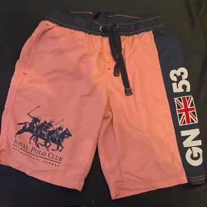 Rosa och marinblå shorts från Royal Polo Club - Snygga rosa shorts med marinblå detaljer från Royal Polo Club. Shortsen har snörning i midjan, tryck med polospelare och brittisk flagga på sidan samt bakficka med logga. Perfekta för sommardagar och sportiga aktiviteter.