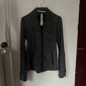 Mörkgrå zip-jacka från Lululemon - Säljer en mörkgrå, figurnära zip-jacka från Lululemon. Jackan har långa ärmar, hög krage och två fickor med dragkedja framtill. Perfekt för träning eller som ett extra lager under kyliga dagar. Storlek S/ liten M. Öppen att diskutera pris!