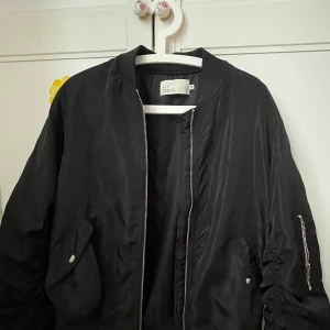 Svart bomberjacka från NLY Trend - Säljer en svart bomberjacka från NLY Trend med klassisk rund krage och dragkedja framtill. Jackan har fickor med tryckknappar och en ärmficka med dragkedja. Perfekt för en cool och avslappnad stil.