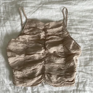 Beige croppad linnetopp med rynkade detaljer - Säljer en beige croppad linnetopp med smala axelband och rynkade detaljer framtill. Toppen har en lätt skrynklig struktur och elastisk baksida för bekväm passform. Perfekt till varma dagar och enkel att matcha med jeans eller kjol.