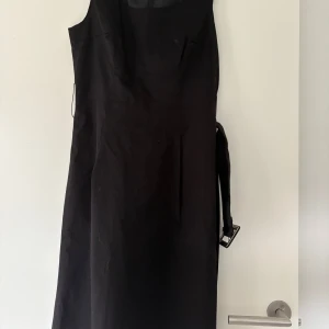 Svart klänning med bälte  - Säljer en svart ärmlös klänning från H&M med markerad midja och matchande bälte. Klänningen har en klassisk, rak passform och är tillverkad i ett slätt tyg. 