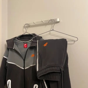 Unlimited nike tech sett!  - Säljer en snygg Nike tech kofta + byxorna till! från Nike i grått och svart med orange logga på bröstet. Tröjan har dragkedja, huva och reflex  detaljer längs ärmar och bröst. Det finns även reflexer över hela setet! Detta paret går inte att hitta i Sverige utan är köpt i Paris. Använd 1 gång. Köpt för 3999kr 