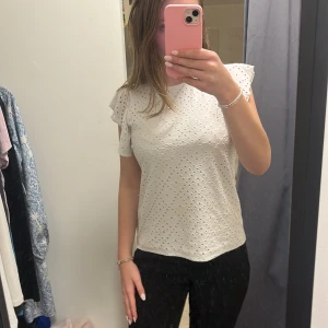 Vit blus med broderade hål och volangärm - Säljer en söt vit blus med broderade hål och korta volangärmar. Blusen har en feminin och luftig känsla, perfekt till vår och sommar. Passar till både jeans och kjol för en fräsch look.