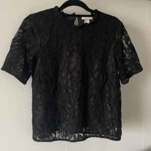 Svart spetsblus med kort ärm - Säljer en svart blus i genombruten spets med blommönster. Blusen har korta ärmar och rund halsringning samt en liten öppning med knapp i nacken. Perfekt för dig som gillar detaljer och vill ha en luftig topp.