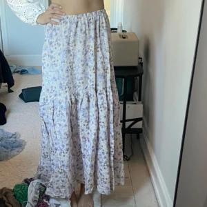 Ljuslila blommig långkjol från H&M - Säljer en ljuslila långkjol från H&M med blommigt mönster och volangdetalj. Kjolen har resår i midjan och är tillverkad i en linnemix som känns luftig och skön. Perfekt för varma dagar och ger en romantisk känsla till outfiten.