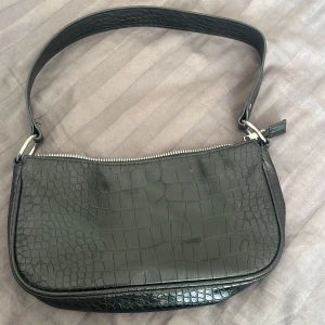 Svart axelväska med krokodilpräglat mönster - Snygg svart axelväska med krokodilpräglat mönster och silvrig dragkedja. Väskan har ett brett axelband och rymligt fack invändigt. Perfekt för att bära dina viktigaste saker med stil.